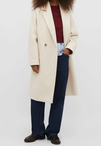 Person iført en cremegul lang frakke, burgunder sweater, lyseblå skjorte, mørkeblå jeans og mørkebrune sko, stående med den ene hånd i lommen.