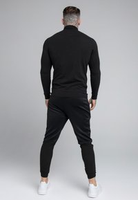 Svart långärmad topp med hög krage och tight passform, parat med svarta joggers med elastiska muddar och vita sneakers.