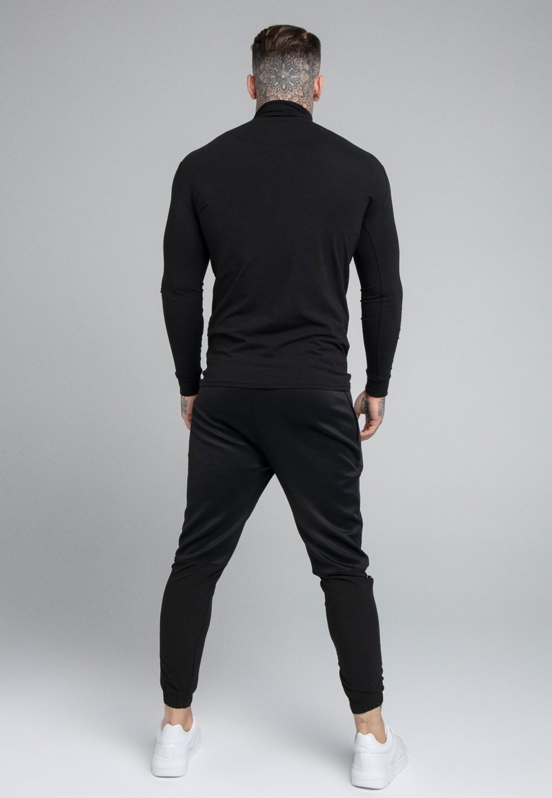 Svart långärmad topp med hög krage och tight passform, parat med svarta joggers med elastiska muddar och vita sneakers.