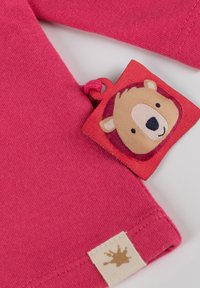 Rosa Baumwollhemd mit einem Bären-Themen-Stoffetikett. Beinhaltet ein strukturiertes, weißes Etikett mit einem braunen Spritzdesign nahe dem Saum.