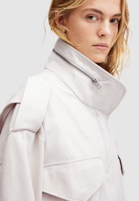 Chaqueta gris claro con un alto cuello, cierre con cremallera y detalles en los hombros en ángulo. Presenta grandes bolsillos laterales y una textura suave.