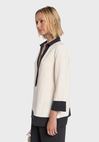 Chemise en lin blanc avec col et poignets noirs, présentant un profond col en V et des manches trois-quarts, texture lisse et lignes géométriques.