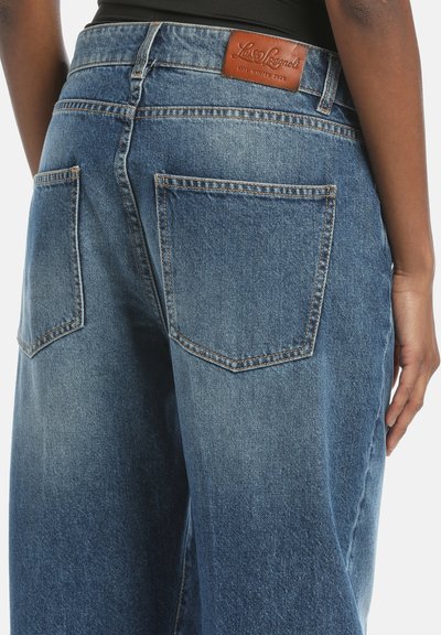 Jeans de mezclilla azul de pierna ancha con cintura alta, que presentan dos bolsillos traseros y un parche de cuero marrón con el logo en la cinturilla.