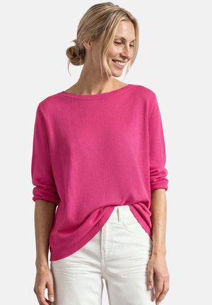 Vrouw met blond haar in een staart, draagt een felroze trui met lange mouwen en een witte broek, glimlachend en naar beneden links kijkend.