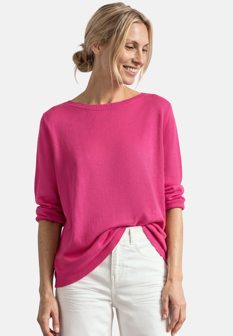 Femme aux cheveux blonds tirés en arrière, portant un pull rose vif à manches longues et un pantalon blanc, souriante et regardant vers le bas à sa gauche.