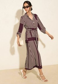Burgunderrotes Kleid mit einem geometrischen cremefarbenen Muster, langen Ärmeln, einem taillierten Bund und einem Kragen. Weiße Riemchensandalen und eine kleine cremefarbene Tasche.