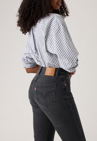 Femeie cu păr creț, purtând o cămașă albă cu dungi albastre, băgată în blugi negri din denim, cu talie înaltă, arătată din spate și lateral.
