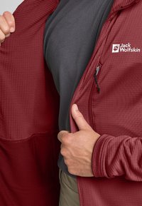 Bordeaux jacket met een getextureerde voorkant, ritssluiting en logo; gedragen over een grijze trui. Opvallende details zijn de gladde en gepatternde stof.