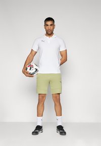 Weißes Poloshirt, olivgrüne Shorts, schwarze Sportschuhe, stehend mit einem Fußball, mit einem Logo auf dem Shirt und den Shorts.