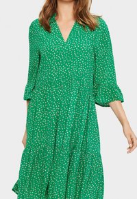 Zelené šaty s výstřihem do V, tříčtvrtečními rukávy, vrstveným designem a bílými puntíky. Vyrobené z lehkého materiálu s vláčnou texturou.
