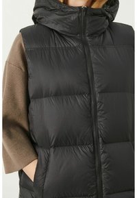 Zwarte gewatteerde mouwloze jas met capuchon, met de ritssluiting omhoog, gedragen over een bruine lange mouw top, hand in de jaszak gestoken.