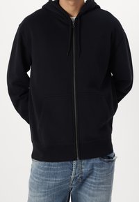 Sudadera negra con cremallera, hecha de tela suave, con bolsillo frontal, capucha ajustable con cordón y puños y bajo acanalados. Llevada sobre unos vaqueros.