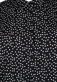 Chemise noire à boutons avec des pois blancs irréguliers et un col, montrant les boutons sur le devant et la texture du tissu en gros plan.