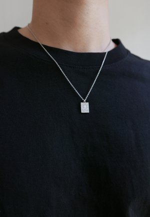 Silver chain necklace features a square pendant with a glittering surface, worn on a black shirt. The chain is delicate and textured.