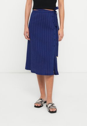 Marineblauer Wickelrock mit weißen vertikalen Streifen, Knopfdetails und gebundenem Taillenband. Kombiniert mit schwarzen und weißen texturierten Slide-Sandalen.