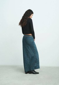 Mujer con cabello largo y oscuro, vistiendo una blusa negra de manga larga, una falda maxi de patrón azul y botas negras hasta el tobillo, de pie contra un fondo sencillo.