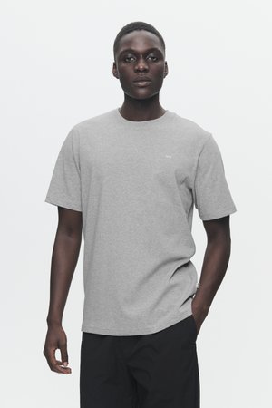 Grå crew-neck t-shirt i bomuld, kortærmet, løs pasform, med minimal branding på brystet. Modellen har sorte bukser på.