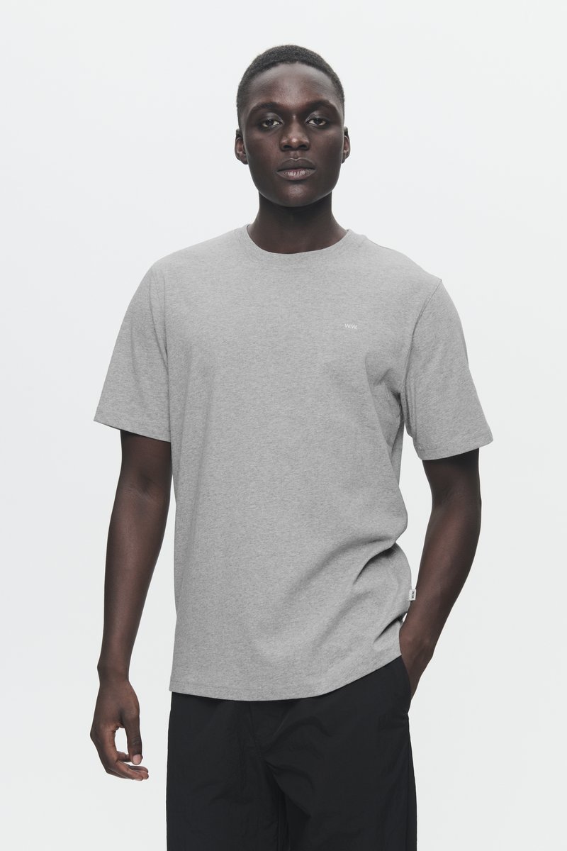Gray crew-neck t-shirt in cotton, short sleeves, loose fit, with minimal branding on the chest. Model wearing black pants.