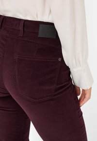 Bordeaux corduroy broek met een gladde textuur, voorzien van een zwart leren label en achterzakken. De stof heeft een subtiel geribbeld patroon.