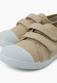 Chaussures en toile beige avec deux sangles Velcro, tissu texturé, forme arrondie et une coque de orteil en caoutchouc blanc texturé.
