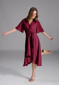 Robe portefeuille bordeaux avec des manches volantées, ourlet asymétrique et taille nouée, associée à des sandales à talons dorées à lanières. Texture de tissu lisse.