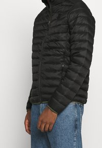 Giacca a vento nera con design trapuntato, colletto alto, chiusura con zip e accenti verdi all'orlo e ai polsini; materiale leggero e strutturato.