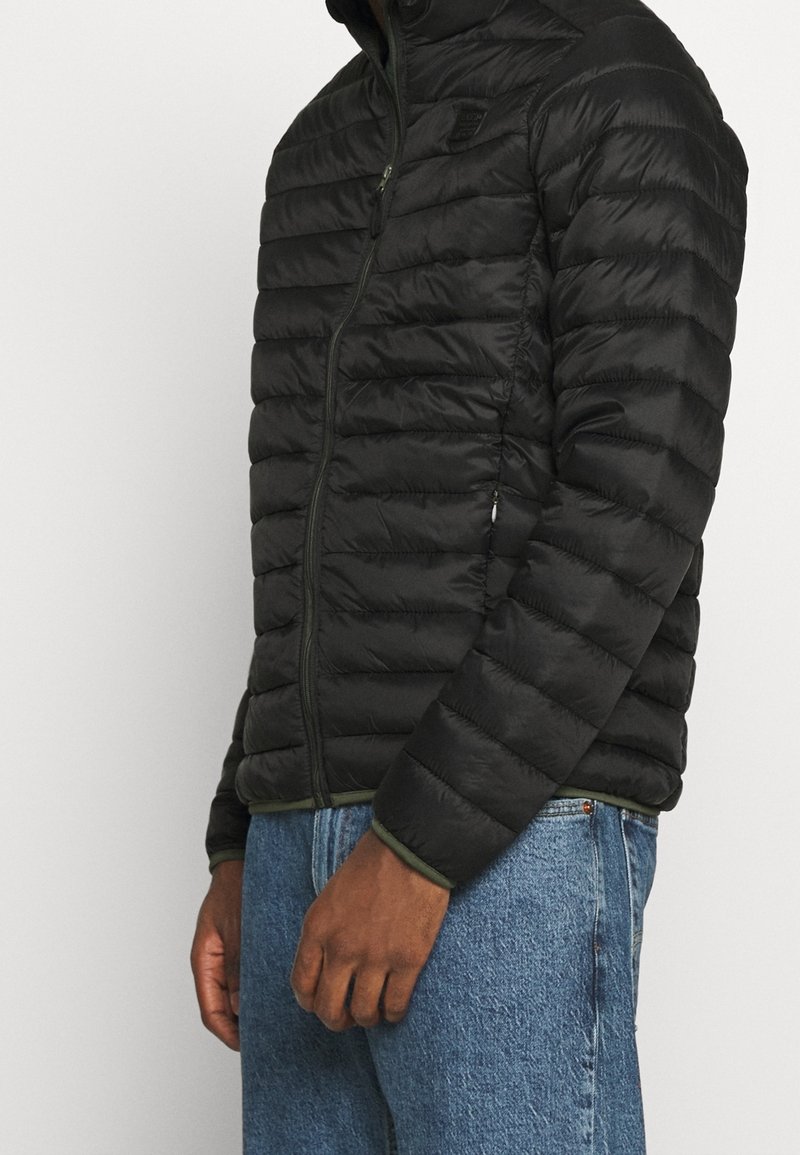 Giacca a vento nera con design trapuntato, colletto alto, chiusura con zip e accenti verdi all'orlo e ai polsini; materiale leggero e strutturato.