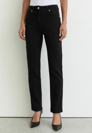 Kvinde iført sorte jeans med lige ben og sorte spidse høje hæle står op ad en hvid væg.
