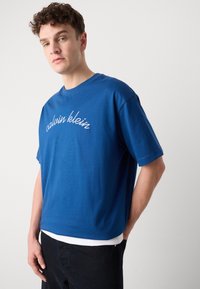 Mėlyna medvilninė marškinėliai trumpomis rankovėmis, su apvaliu kaklu ir balta siuvinėta "calvin klein" logotipu per krūtinę.