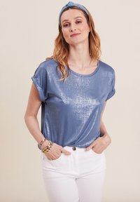 Glanzend blauw T-shirt met korte mouwen en een ronde halslijn, gecombineerd met witte broeken. Accessoires zijn kleurrijke armbanden en een blauwe haarband.