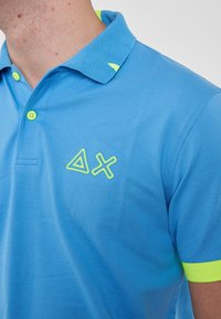 Polo shirt azzurro chiaro realizzato in tessuto testurizzato, con colletto in evidenza verde neon e logo giallo neon sul petto.