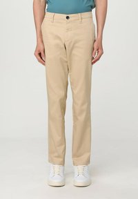 Pantaloni beige in tessuto liscio, con chiusura frontale a bottone, due tasche laterali e design slim fit. Indossati con sneaker bianche.