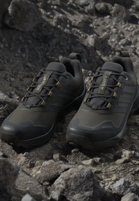 Chaussures de randonnée Adidas Terrex noires avec lacets sur terrain rocheux, montrant un design durable et une utilisation robuste en extérieur.