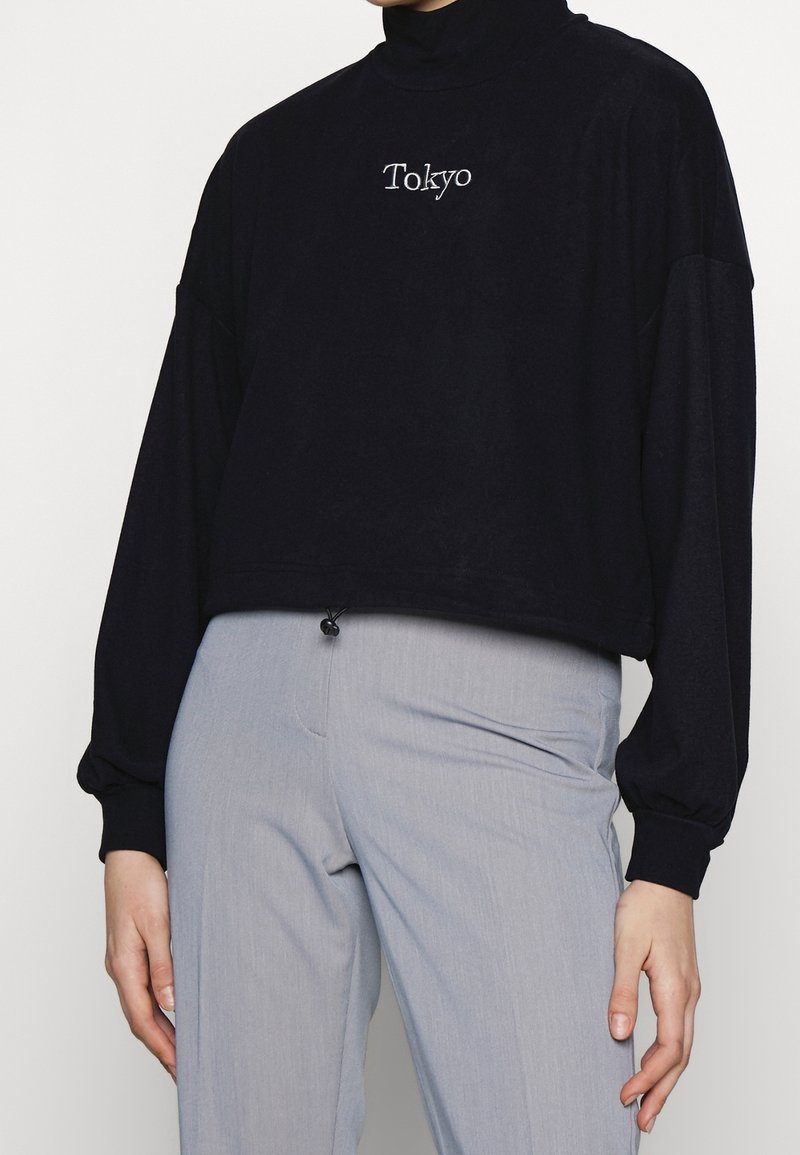 Persoon draagt een zwarte coltrui met lange mouwen en de tekst "Tokyo", en lichtgrijze broek met hoge taille, handen ontspannen langs het lichaam.