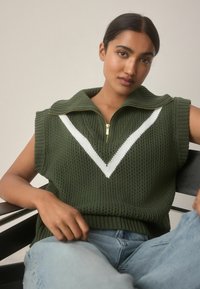 Ärmelloser, dunkelgrüner Strickpullover mit hohem Kragen und einem weißen V-Muster. Verfügt über einen Frontreißverschluss und eine gerippte Textur am Saum.