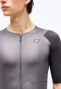 Κοντινό πλάνο ενός ατόμου που φοράει γκρι αθλητική μπλούζα με φερμουάρ και ένα μικρό οβάλ λογότυπο στο στήθος, με υφασμένα μαύρα κοντά μανίκια.