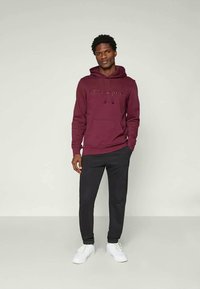 Bordeauxrode hoodie met een voorzak en een trekkoord capuchon, met het "Champion" logo in een subtiele, donkere tint. Gecombineerd met zwarte joggingbroek en witte sneakers.
