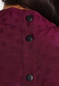 Haut en tissu bordeaux avec un motif à pois texturé, comportant une rangée de boutons noirs au dos. Texture lisse et design ajusté.