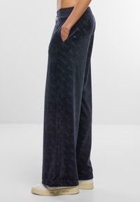 Navy velour wide-leg pants feature side pockets, an elastic waistband, and an all-over logo pattern. Paired with white sneakers.