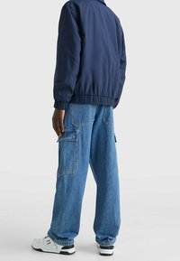 Navy bomber jacket with elastic waistband, paired with light blue cargo jeans featuring multiple pockets and a relaxed fit. White sneakers.