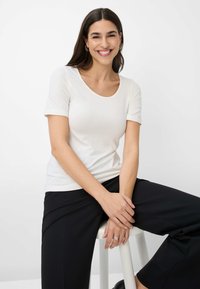 Witte T-shirt met korte mouwen en een ronde hals, gemaakt van soepele stof, gecombineerd met zwarte broek. Model zittend op een kruk, met de handen gekruist.