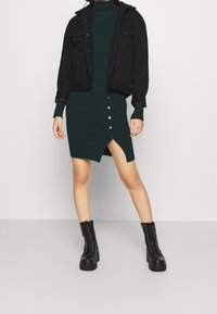 Svart denimjacka med shearlingfoder, grön ribbad klänning med framknappar och sidöppning, i kombination med svarta snörstövlar.