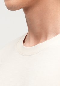 Světlé béžové tričko s žebrovaným kulatým výstřihem a hladkým, mírně texturovaným materiálem. Detail na výstřih a horní část trupu.