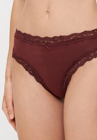 Burgunderfargede truser med blondekant, med en glatt kropp, og med en flat sløyfedetalj foran samt delikate blonder langs kantene.