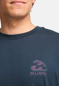 Camiseta de algodón azul marino con un logo de olas en verde y rosa, acompañado del texto "BILLABONG ADVENTURE DIVISION" debajo del gráfico.