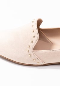 Chaussure slip-on en daim beige avec un design festonné et des clous dorés le long du bord supérieur. Texture lisse et forme de bout arrondi.