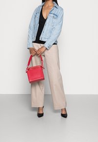 Sac à main en cuir rouge de forme carrée, avec une poignée courte et des détails de couture. Porté avec une veste en jean claire et un pantalon beige à jambes larges.