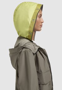 Veste légère olive avec une capuche structurée en vert fluo. Possède une fermeture éclair à l'avant et un cordon de serrage ajustable au niveau de la capuche.
