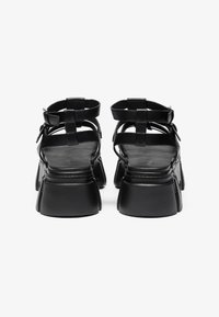 Black leather sandals with a chunky heel, featuring strap details and an open back design. The heel has a textured, matte finish.