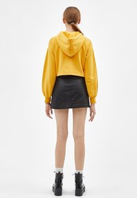 Mujer de pie de espaldas, vistiendo un sudadera amarilla corta, una falda mini de cuero negra, calcetines blancos y botas de plataforma negras con cordones.